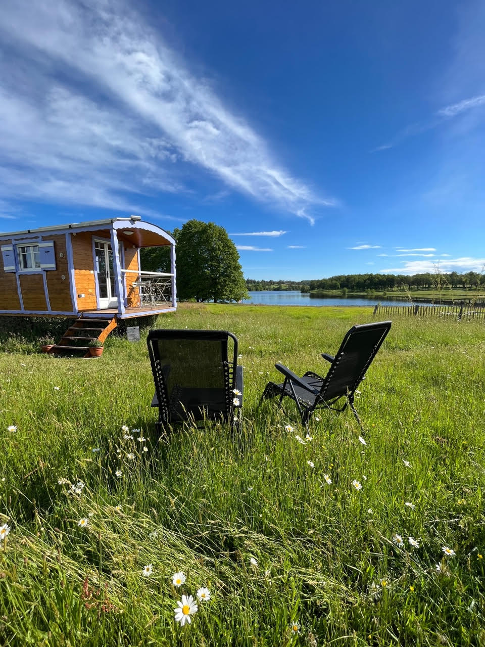 A vintage caravan is positioned beside a calm lake, surrounded by lush green grass. Two folding chairs are placed in the foreground, facing the water. The sky is clear with scattered clouds, creating a tranquil outdoor atmosphere.