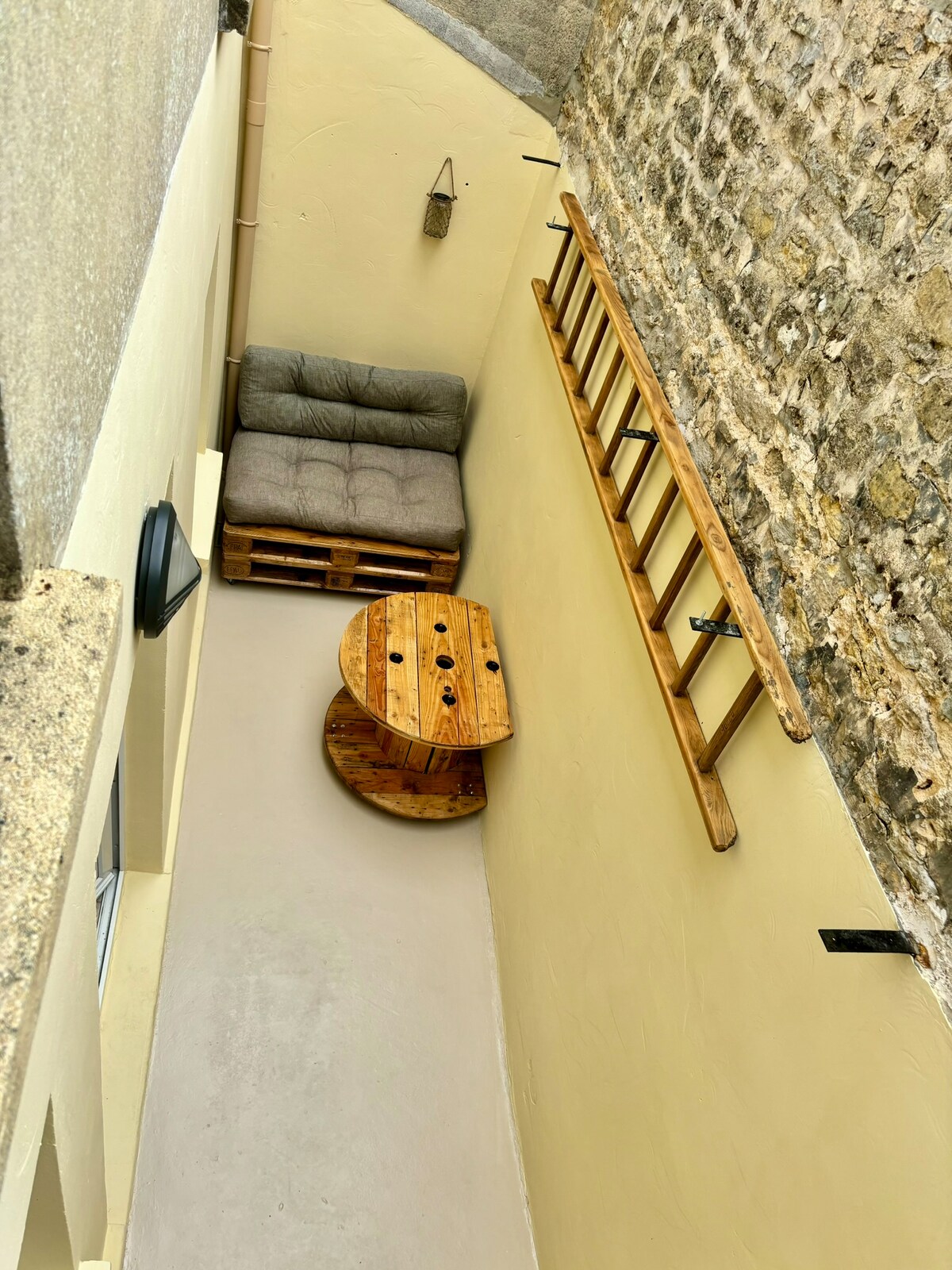 An intimate courtyard features a cozy seating arrangement consisting of a palette sofa and a wooden round table. The surrounding stone walls add character to the space, while soft lighting emphasizes a relaxing atmosphere perfect for outdoor enjoyment.