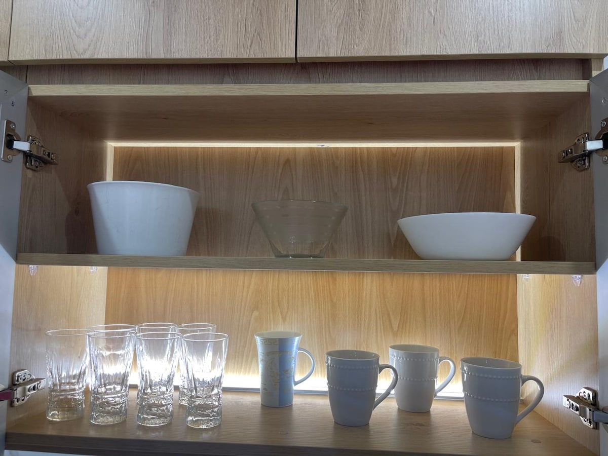 A wooden cabinet interior is illuminated softly, displaying a set of glassware including several clear glasses and mugs. Two white bowls and a glass serving dish are positioned on the upper shelf, creating a functional and organized kitchen space.