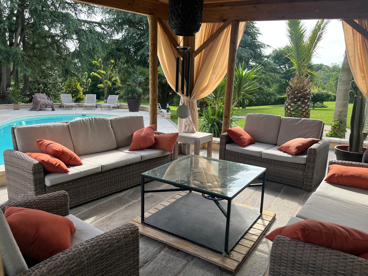 A covered outdoor seating area is presented, featuring multiple comfortable sofas adorned with cushions. A glass table is centered among the seating. A pool glistens in the background, surrounded by lush greenery and landscaped gardens.