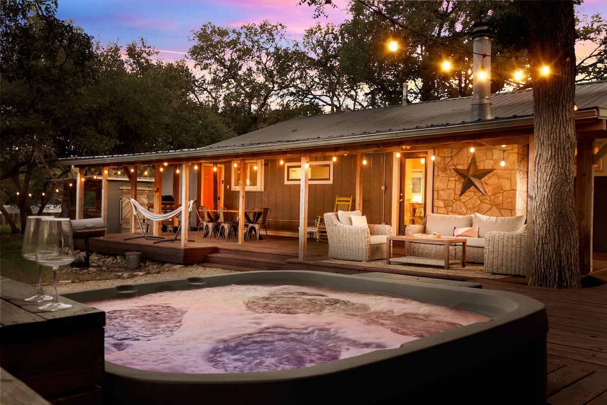 A hot tub sits in the foreground, with illuminated bubbles creating a soothing ambiance. The main home features a covered patio, adorned with string lights, designer furniture, and wooden accents, providing a cozy gathering space amid a tranquil, rustic setting.