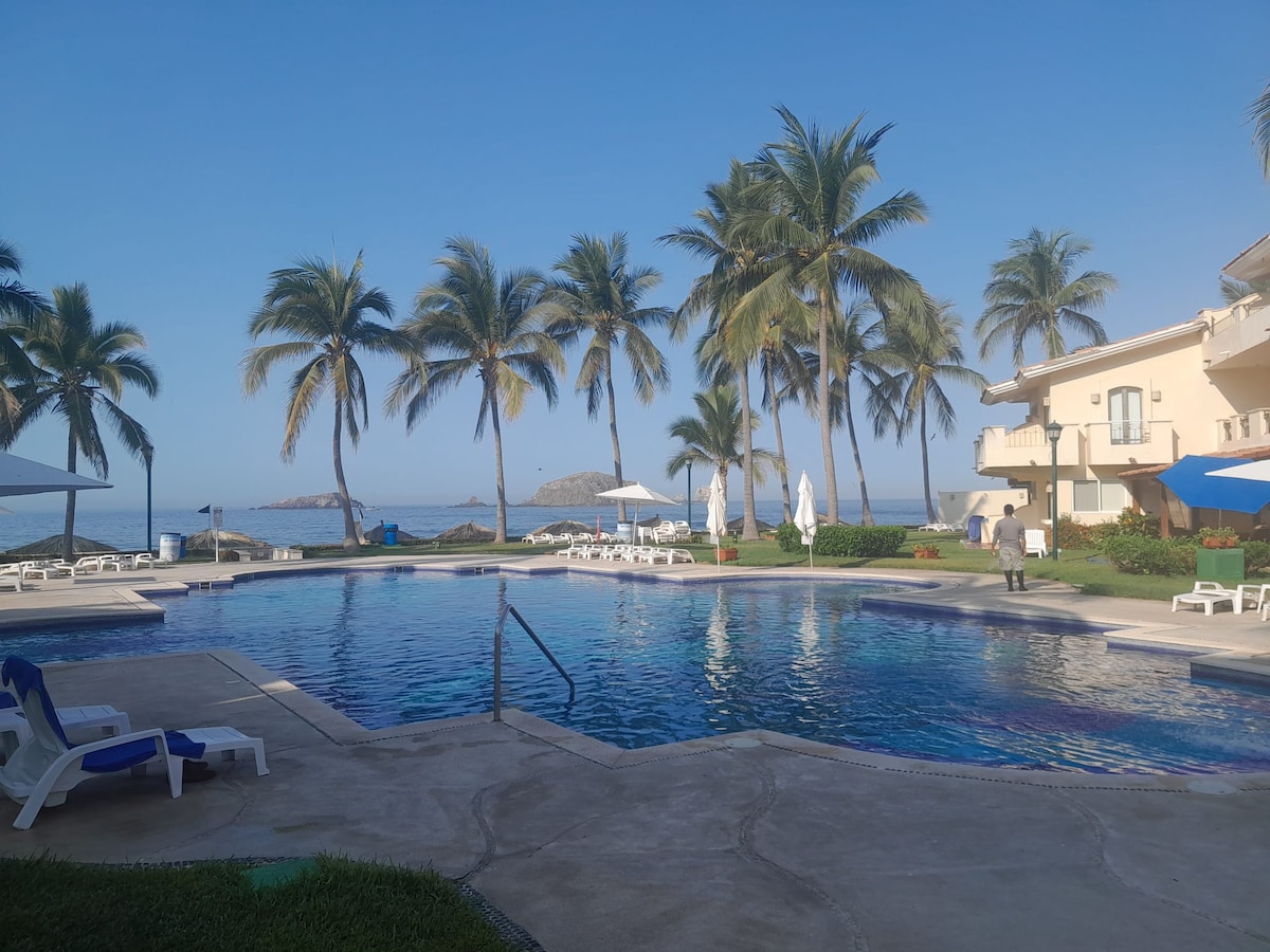 The image depicts a serene pool area surrounded by palm trees and overlooking the ocean. Lounge chairs are arranged around the pool, and umbrellas provide shade. The clear blue water of the pool reflects the bright sky, creating a calm atmosphere.