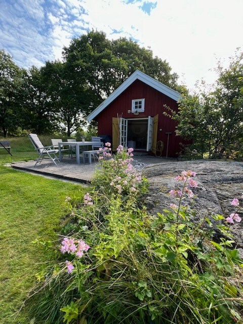 A charming red cottage is nestled in a natural setting, surrounded by greenery and wildflowers. Outdoor furniture is arranged on a stone patio, inviting relaxation. A dirt path leads to the entrance, framed by lush grass and trees in the background.