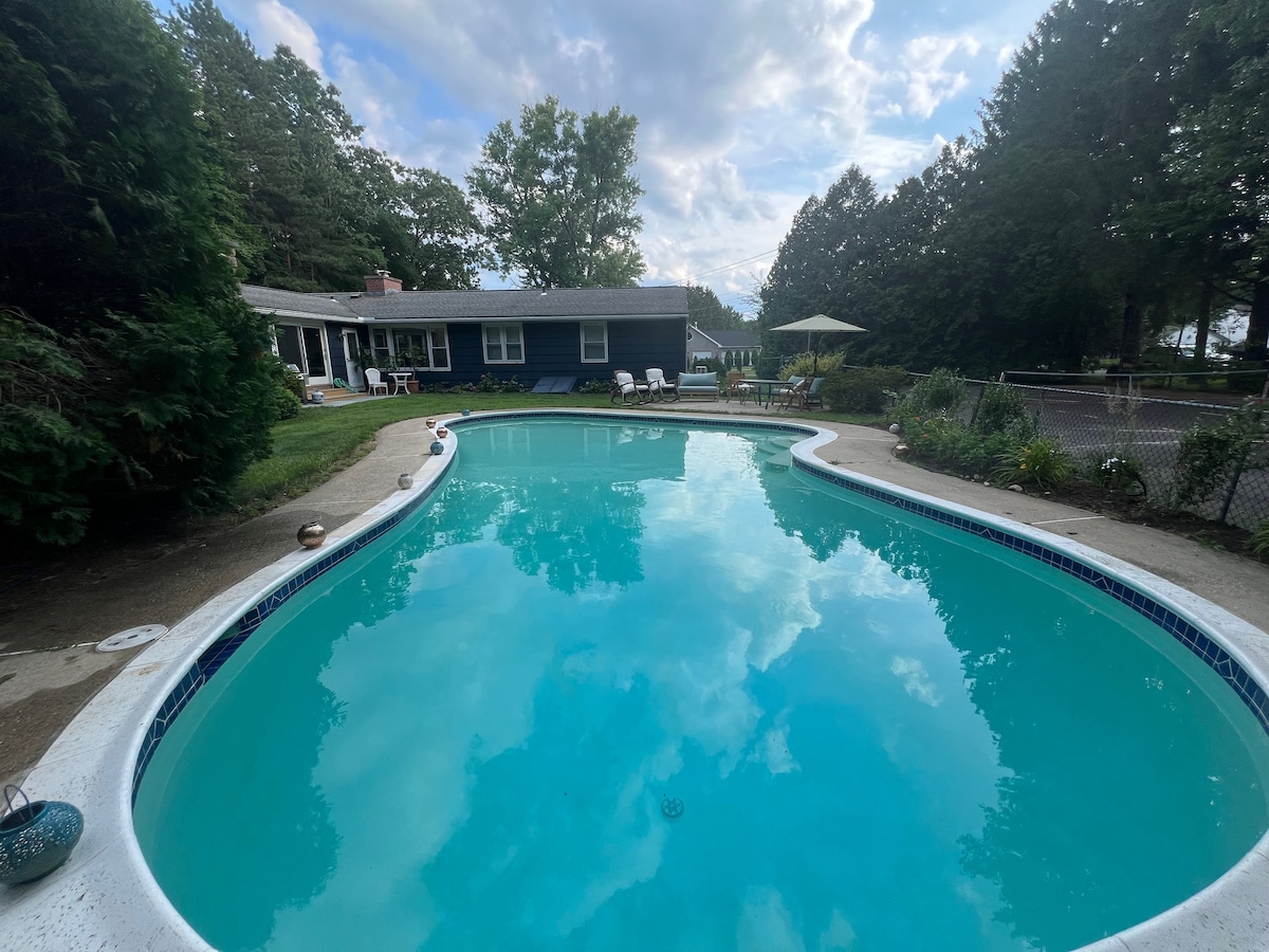 A serene outdoor pool is framed by lush greenery and a well-maintained yard. Relaxing seating options can be seen around the pool area, while a sun umbrella provides shaded comfort. The inviting turquoise water reflects the sky and surrounding trees.
