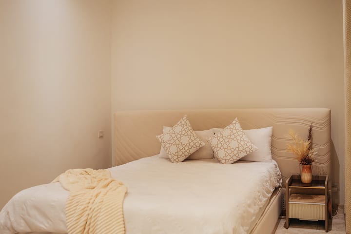 На мала ноќна масичка има цветен аранжман во вазна, додавајќи малку природен декор. Ѕидовите се насликани во топло беж, што придонесува за ведрата и привлечна атмосфера.