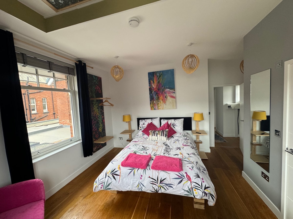 A bright bedroom is featured, furnished with a spacious bed adorned with a floral patterned duvet. Two bedside lamps provide illumination, while art hangs on the walls. A window allows natural light to fill the room, with a view of the street and a small seating area visible.