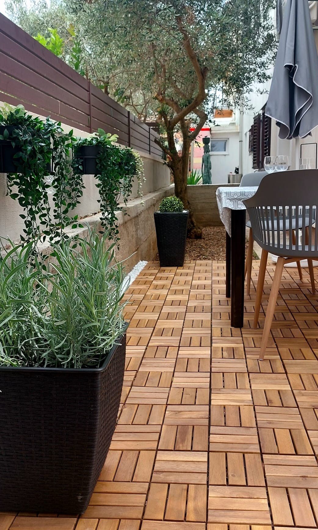 A private outdoor terrace is presented, featuring a rich wooden flooring pattern. Potted plants line the space, adding greenery. A table and chairs are positioned for dining, shaded by an umbrella. A tree provides a natural touch, enhancing the tranquil setting.
