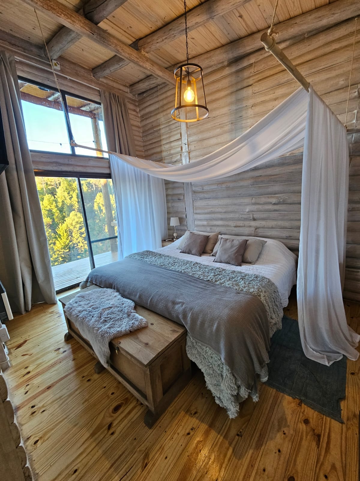 A welcoming bedroom features a spacious bed dressed in soft linens and accented with decorative pillows. Natural light floods the room through large windows, revealing views of surrounding trees. A cozy blanket rests at the foot of the bed, and wooden flooring enhances the rustic feel.
