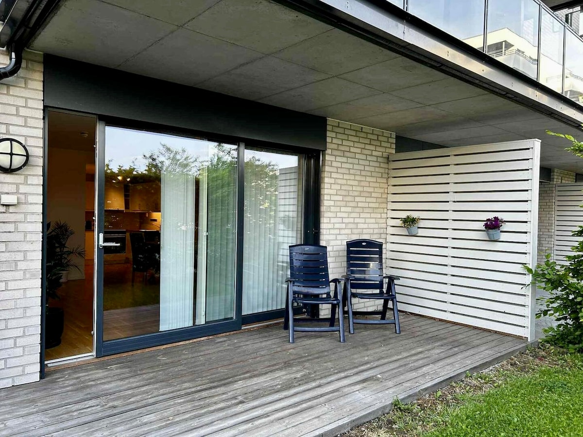 A private balcony features two dark blue chairs on a wooden deck. The sliding glass doors provide access to the interior, revealing a glimpse of the open-plan living space. Potted plants are mounted on the wall, enhancing the area with natural green elements.