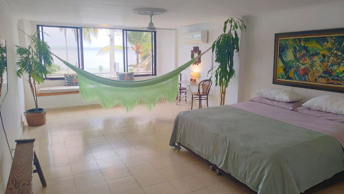A spacious room features a light green hammock suspended between two windows, with views of the beach and palm trees. A comfortable bed with soft bedding occupies one corner, while a small table and chairs add accessible seating options. Natural light fills the space through the large windows.
