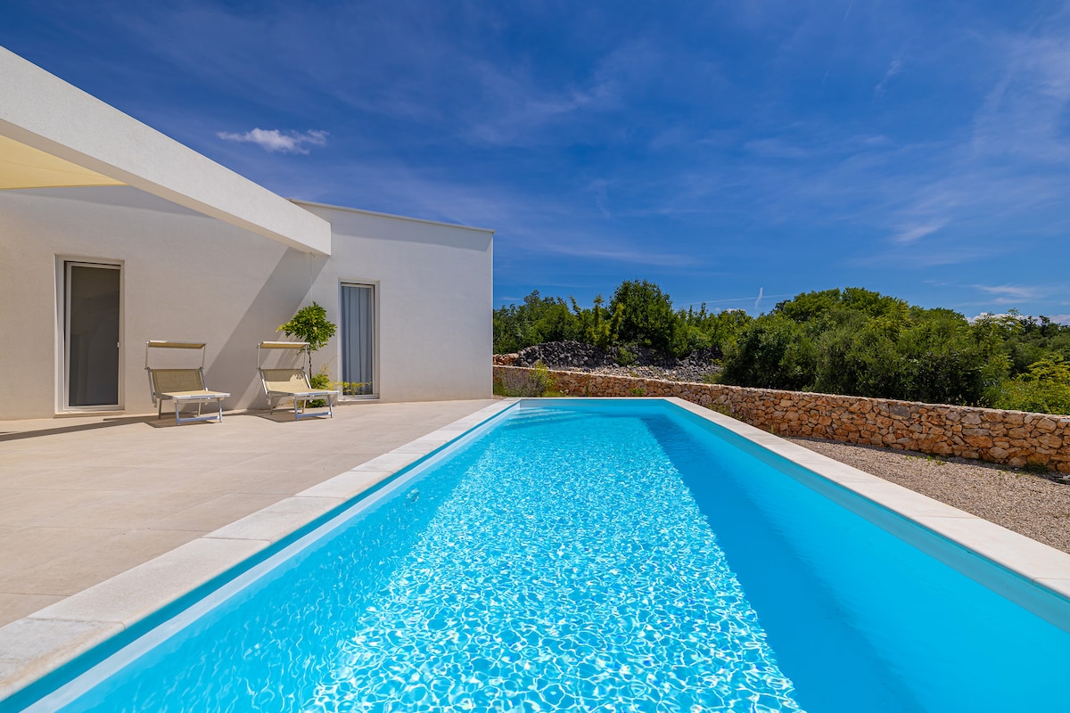 A tranquil pool area is highlighted, with clear blue water reflecting the sky. Sun loungers are positioned beside the pool, surrounded by natural greenery and a stone boundary wall. The structure presents clean lines and a light-colored exterior, contributing to a serene outdoor setting.
