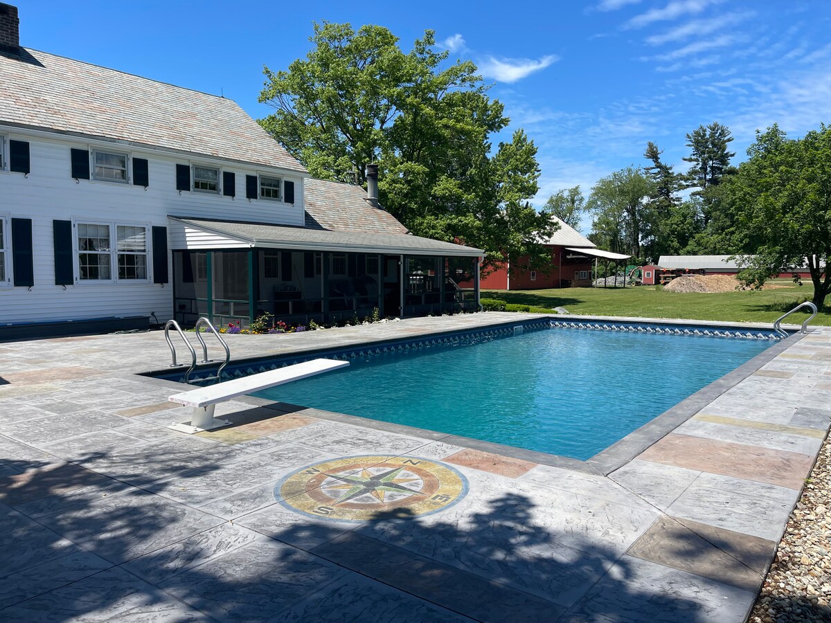 A spacious outdoor swimming pool is surrounded by a concrete deck featuring a decorative compass design. Sunlight glistens on the water's surface, and a diving board is positioned at one end of the pool. The farmhouse and lush greenery form a serene backdrop.
