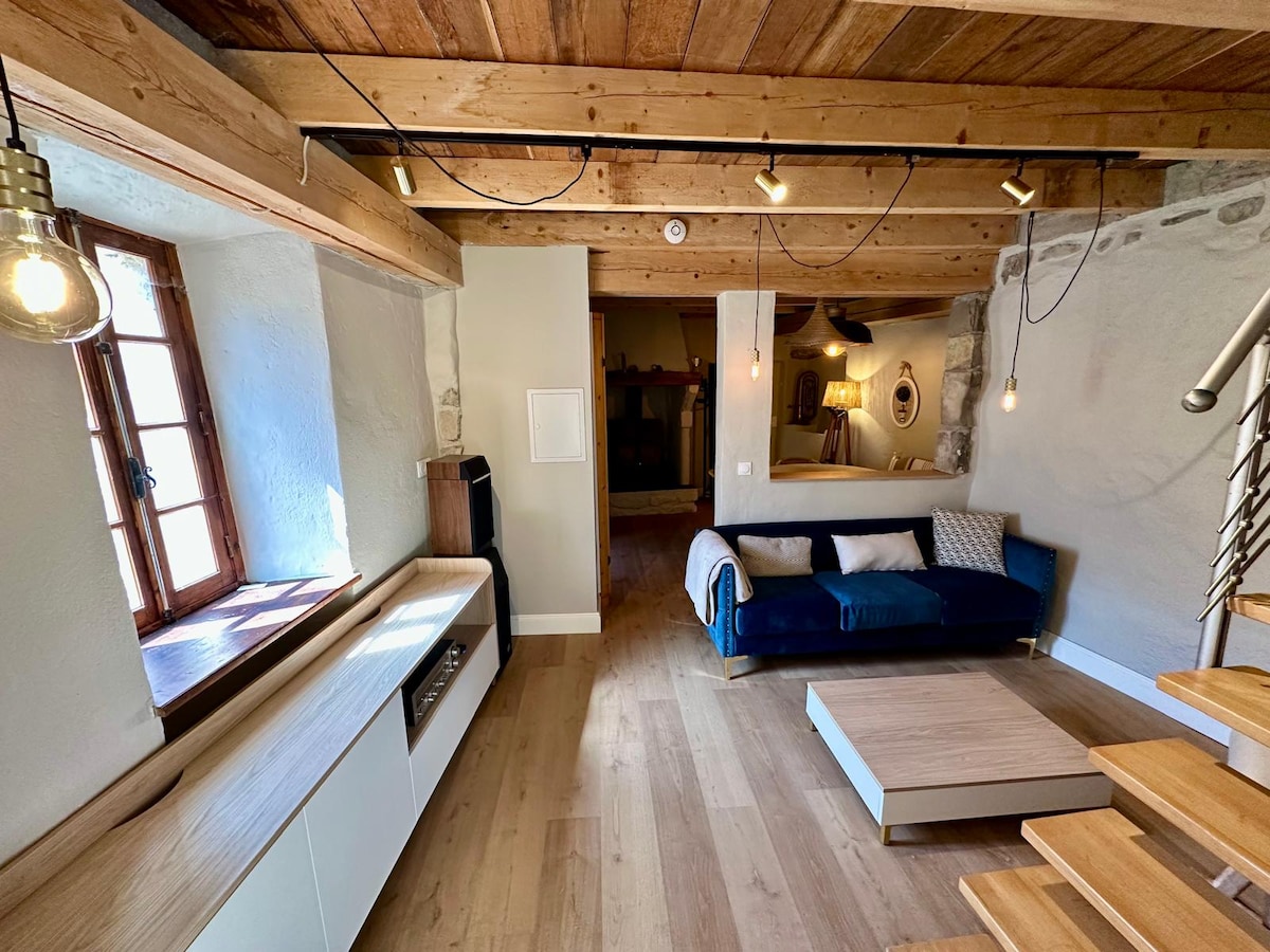 A spacious living area showcases wooden beams overhead and warm tones throughout. A blue sofa is positioned near a large window, allowing natural light to fill the room. Simple decor elements provide a comfortable and relaxed ambiance. A staircase is visible leading to the upper level.