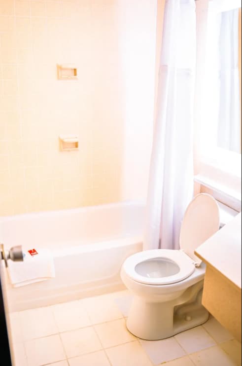 A clean and simple bathroom features a white bathtub with a shower curtain and beige tiles. A toilet is positioned beside a countertop where a towel is neatly placed. Natural light enters through a nearby window.