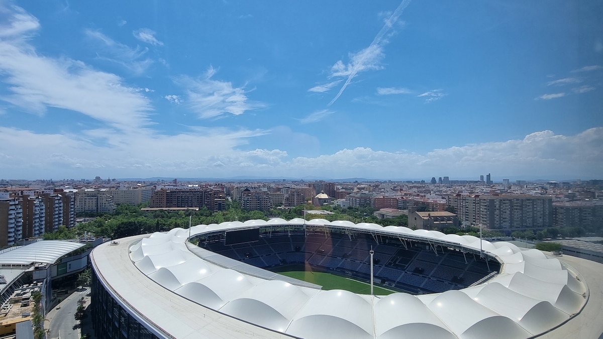 A panoramic view showcases the vibrant city skyline, highlighting numerous buildings against a clear blue sky. In the foreground, a spacious stadium with a distinctive curved roof is visible, surrounded by city greenery and urban developments.
