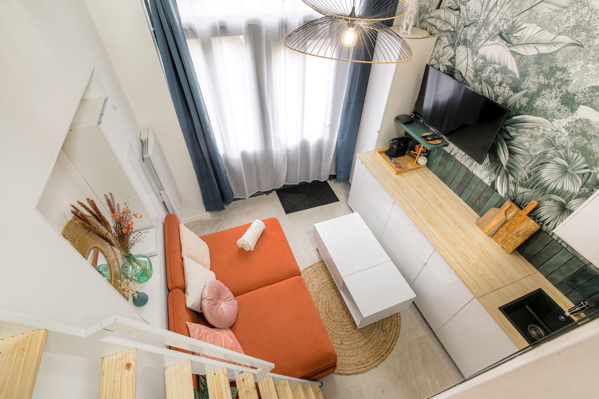 A spacious living area features a bright orange sofa complemented by decorative cushions. Large windows allow natural light to fill the space, while a wall adorned with tropical wallpaper adds a touch of style. A modern TV and a minimalist console table complete the setting.