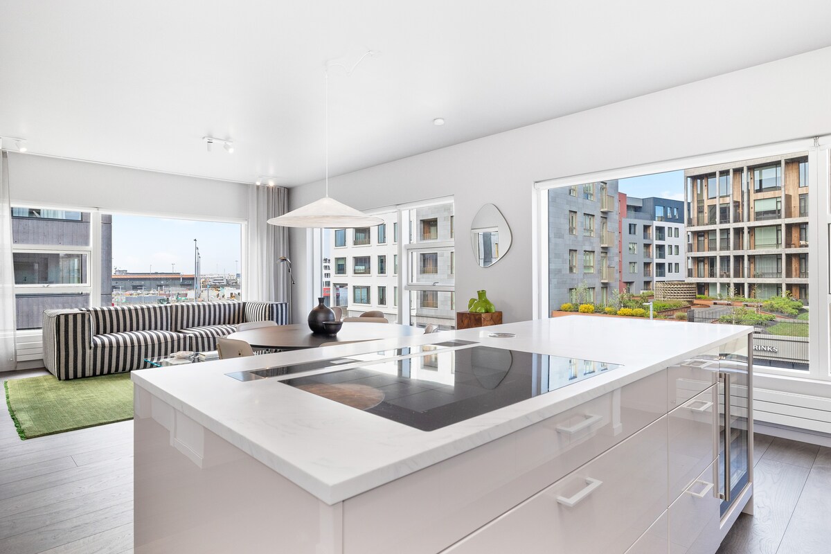 A modern kitchen area features an island with a stovetop and sleek countertops. Large windows provide natural light and offer views of the outdoor space, which is complemented by attractive landscaping. A dining area with a striped sofa and a green rug is visible in the background.