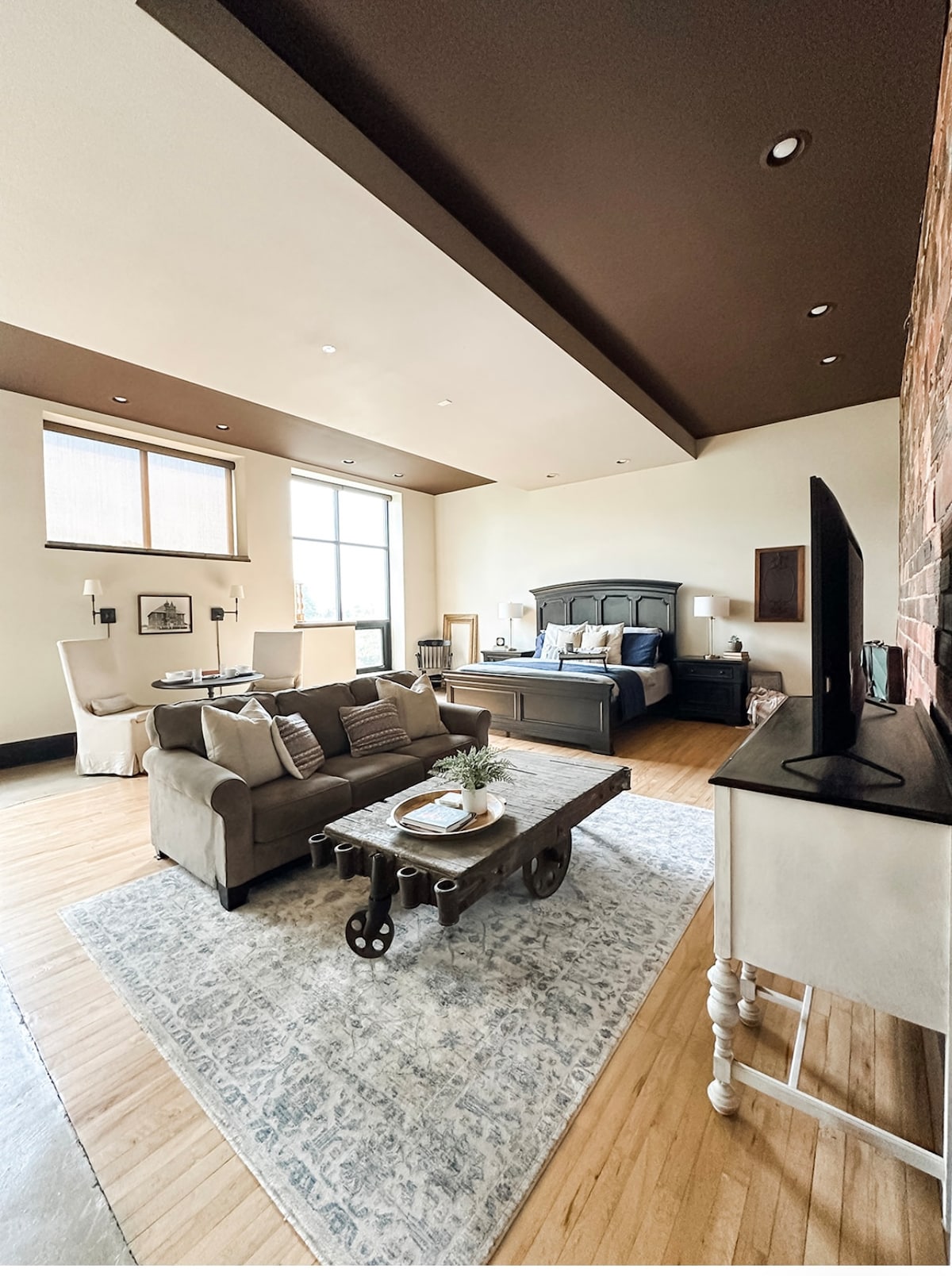 A spacious suite features a comfortable sitting area with a beige couch and rustic coffee table. Natural light is welcomed through large windows, highlighting the warm wood floors and contrasting ceiling. The sleeping area includes a king-sized bed against a dark accent wall, providing a cozy atmosphere.