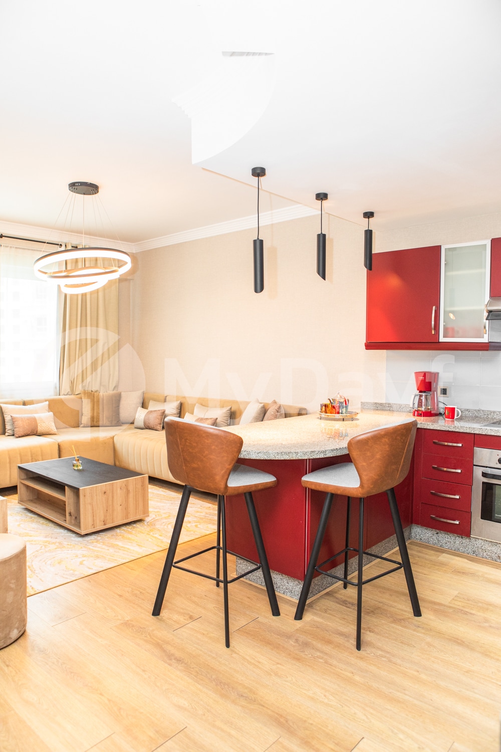 A modern kitchen area is highlighted by a central island with two high stools, red cabinetry, and stainless steel appliances. A cozy seating arrangement is set nearby on neutral-toned fabrics, and natural light filters in through a large window.