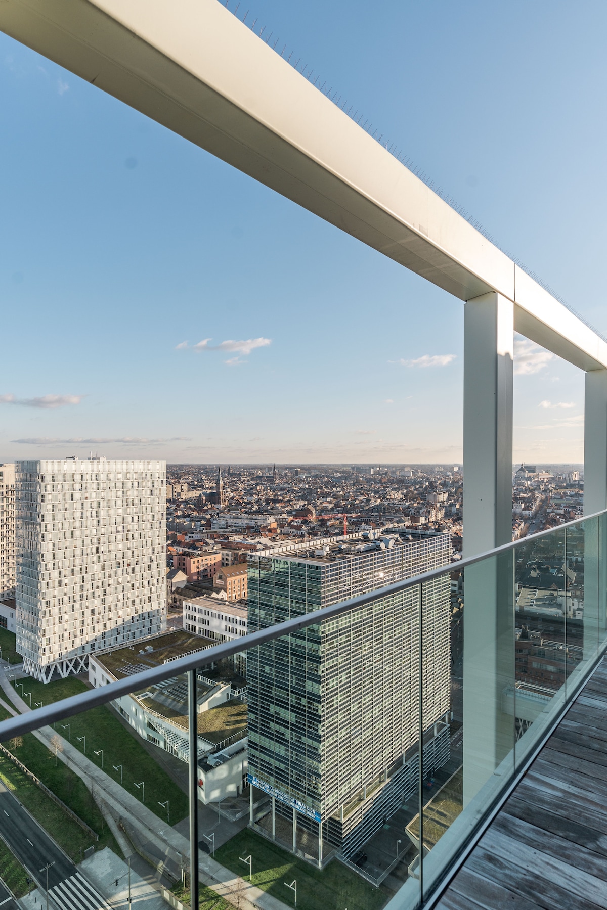 A sweeping view of the city skyline is presented from the private terrace. Modern buildings of various heights dominate the landscape, framed by clear skies and distant horizons. The railing features sleek glass panels, enhancing the unobstructed perspective of the vibrant urban environment below.