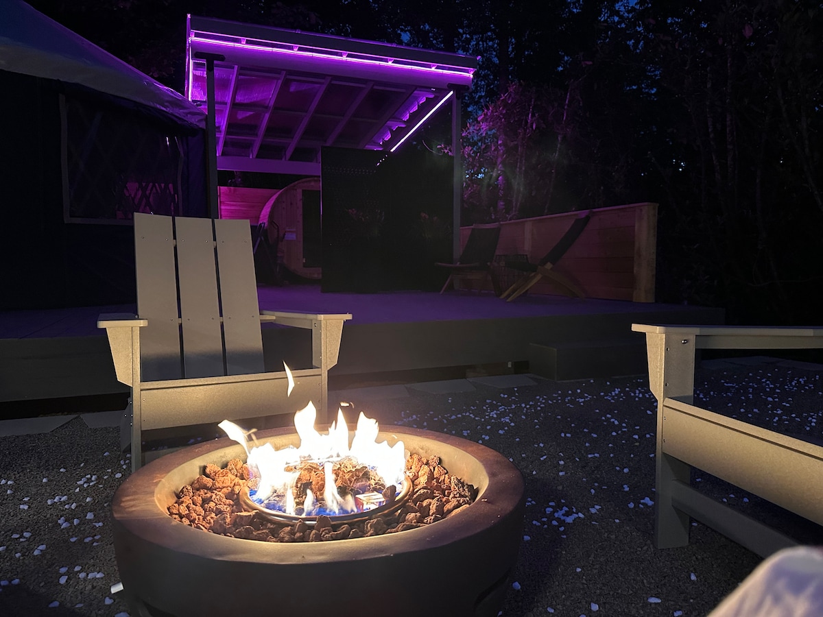 A cozy fire pit is surrounded by two light-colored adirondack chairs, with flames flickering above a bed of natural rocks. In the background, soft purple lighting highlights a covered outdoor space, enhancing the serene ambiance of the surrounding nature.