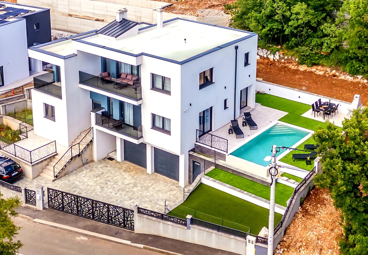 The modern villa is viewed from above, showcasing multiple levels and a sleek exterior. A private infinity pool extends from the terrace, flanked by lounge chairs. Well-manicured lawns and a spacious outdoor dining area are visible, contributing to the expansive setting.