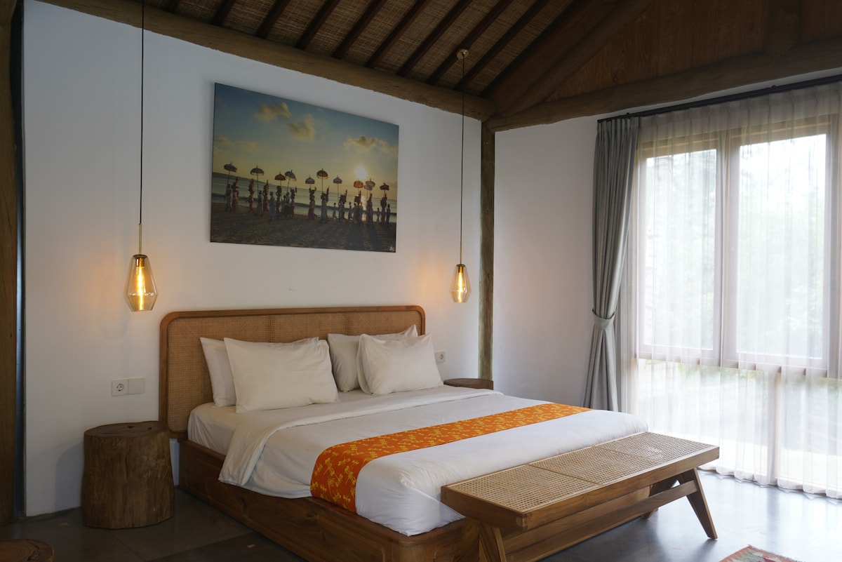 A spacious room features a large bed adorned with neutral-toned linens and an orange decorative accent. Two pendant lights hang above the bed, and a piece of wall art depicting a beach scene adds visual interest. Large windows allow natural light to fill the space.