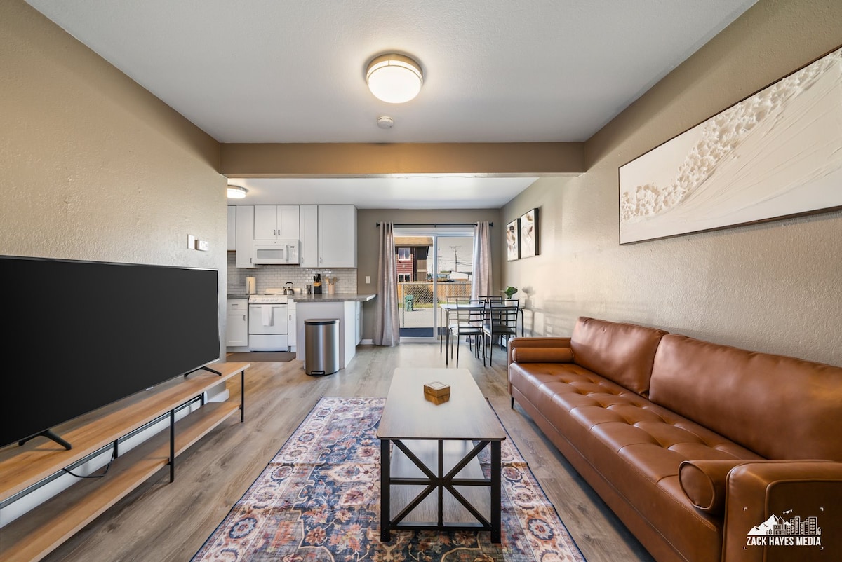 The living area features a leather sofa facing a TV on a low stand. A wooden coffee table sits in front, complementing the patterned area rug. The kitchen is visible through an open layout, with a dining table and chairs beside the glass door leading to the outdoor space.