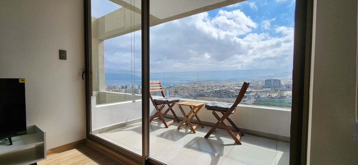 A balcony is featured with two wooden chairs and a small table, offering expansive views of the surrounding landscape. Glass doors allow natural light to enter the interior space, enhancing the sense of openness while framing the scenic vista.