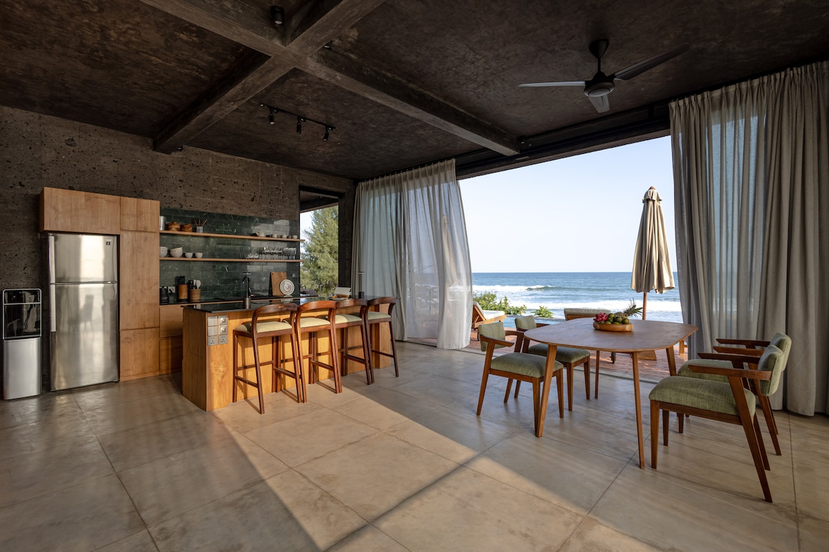 An open-concept dining area features a wooden table surrounded by chairs, with a view of the ocean through large glass doors. A well-equipped kitchen is visible, complemented by a refrigerator and minimalistic decor. Light pours in from expansive windows, creating a bright and airy atmosphere.