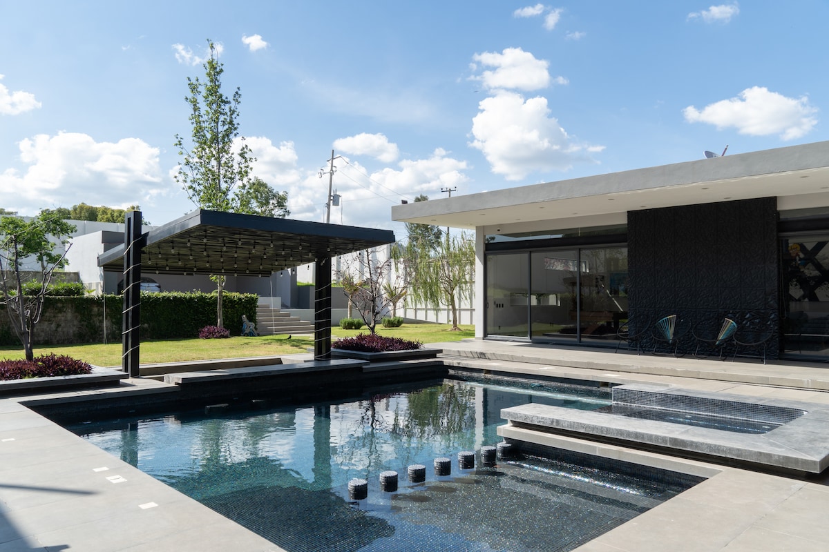 A modern outdoor space features a sleek pool with a dark tiled surface, surrounded by organized landscaping. A shaded pergola stands to one side, offering seating. Large glass doors lead into the interior, showcasing natural light and open design elements.