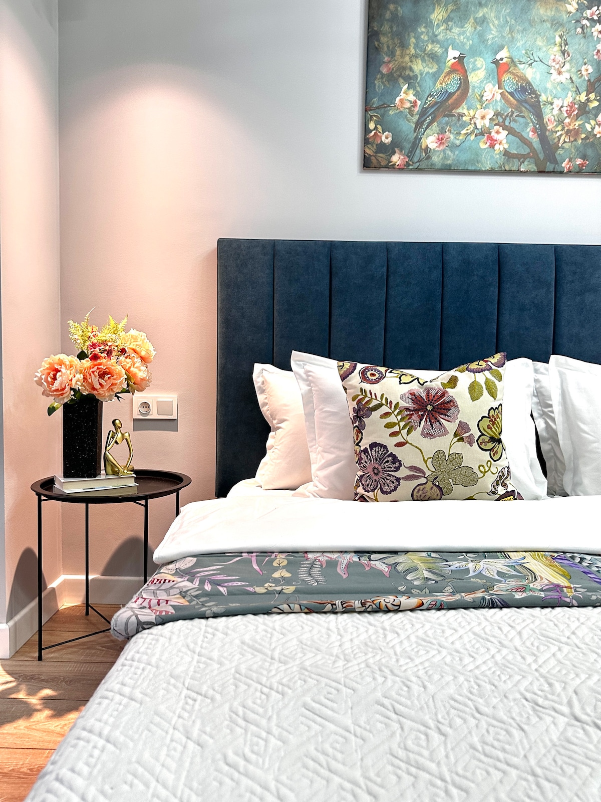 Un dormitorio con una cama con cabecera azul crea comodidad y comodidad. En la mesita de noche hay un arreglo floral y elementos decorativos, y encima de la cama cuelga una imagen de aves exóticas, agregando arte y calidez al espacio.