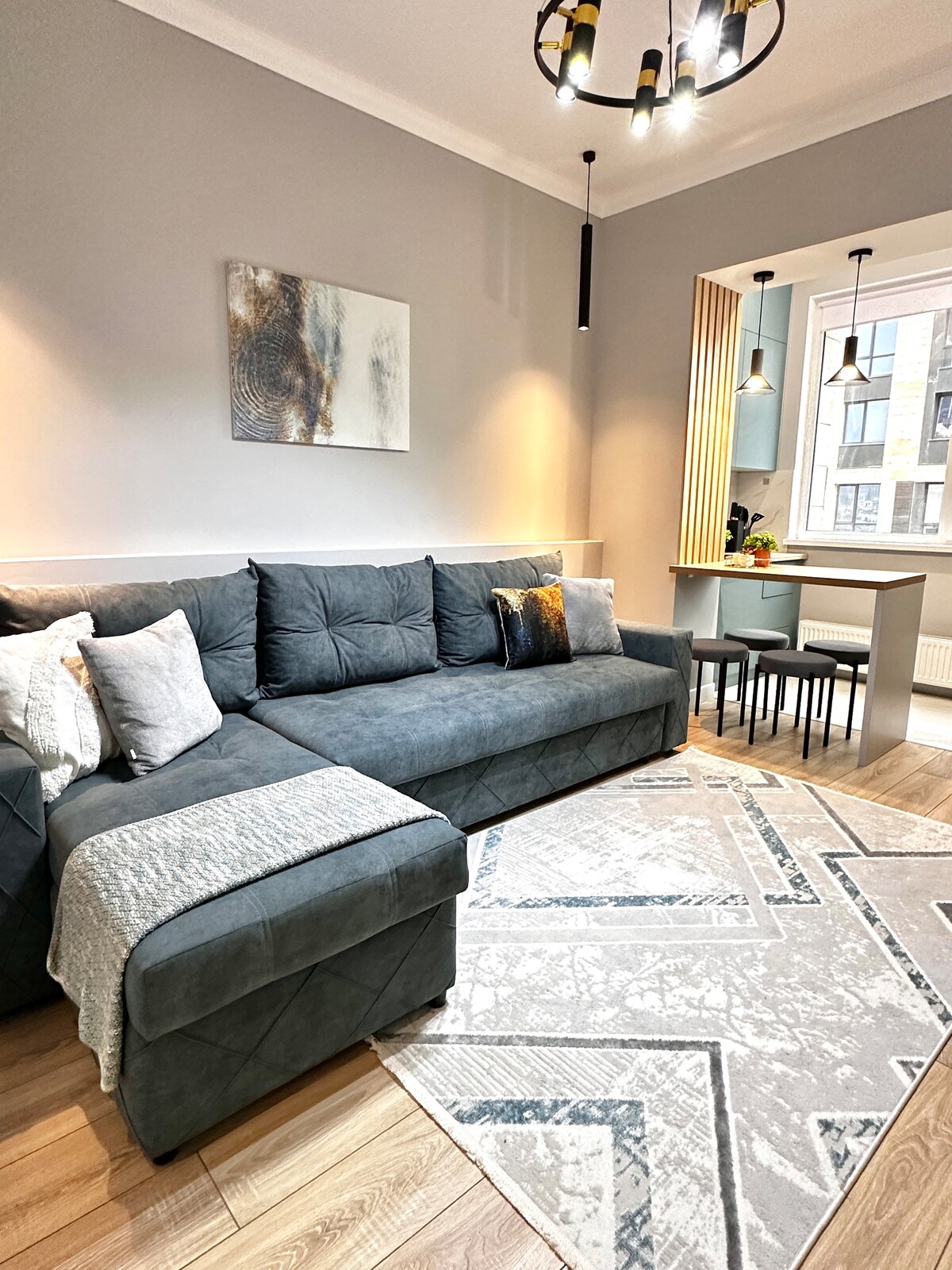 La foto muestra una acogedora zona de estar con un sofá gris suave decorado con almohadas decorativas, así como una alfombra ligera y geométrica, que le da al espacio un aspecto fresco y elegante.