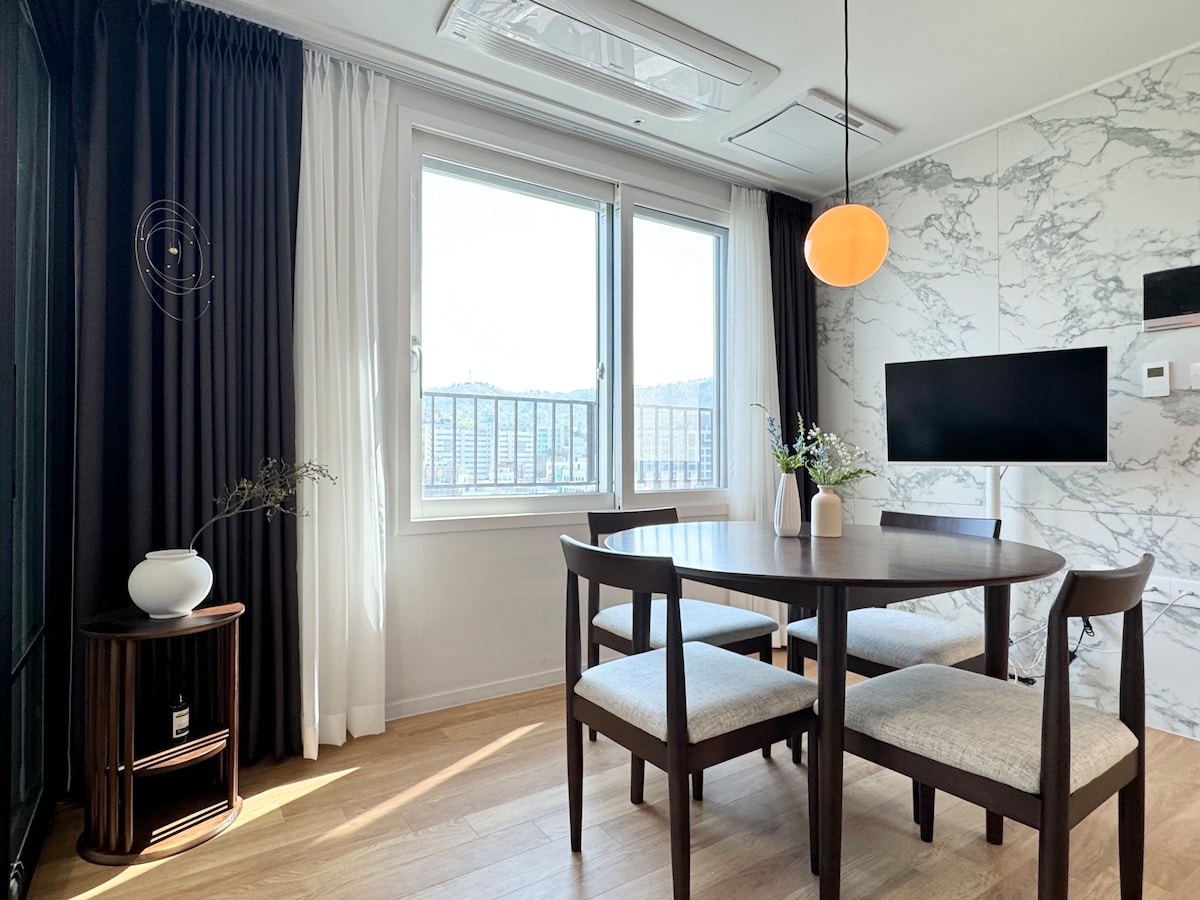 A dining area features a round table surrounded by four light-colored chairs. Natural light filters through a large window adorned with dark curtains and white sheers. A decorative vase with greenery rests on the table, while a wall-mounted TV is visible nearby.