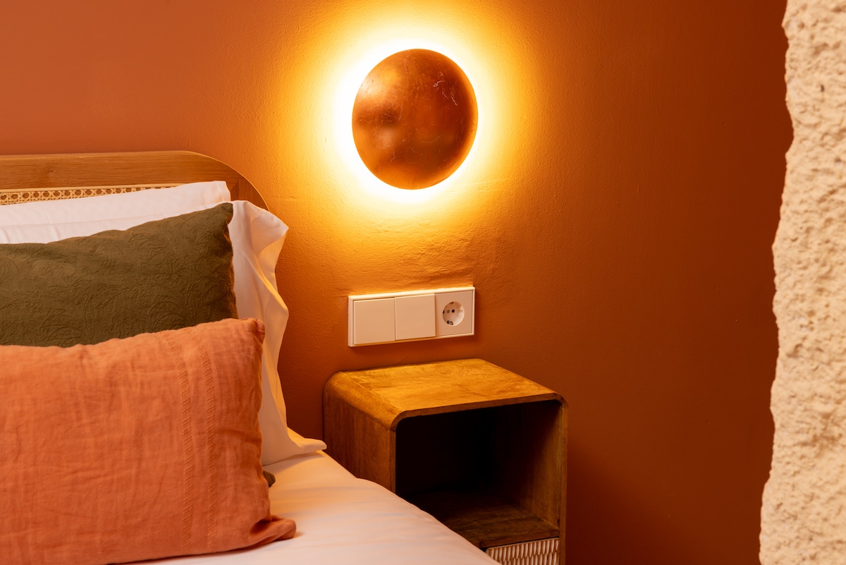 The inviting sleeping area is adorned with earthy tones, featuring a spacious bed with layered cushions. A warm light fixture above creates a cozy ambiance, while a bedside table complements the decor. The wall is painted in a soft terracotta hue, enhancing the room's overall comfort.