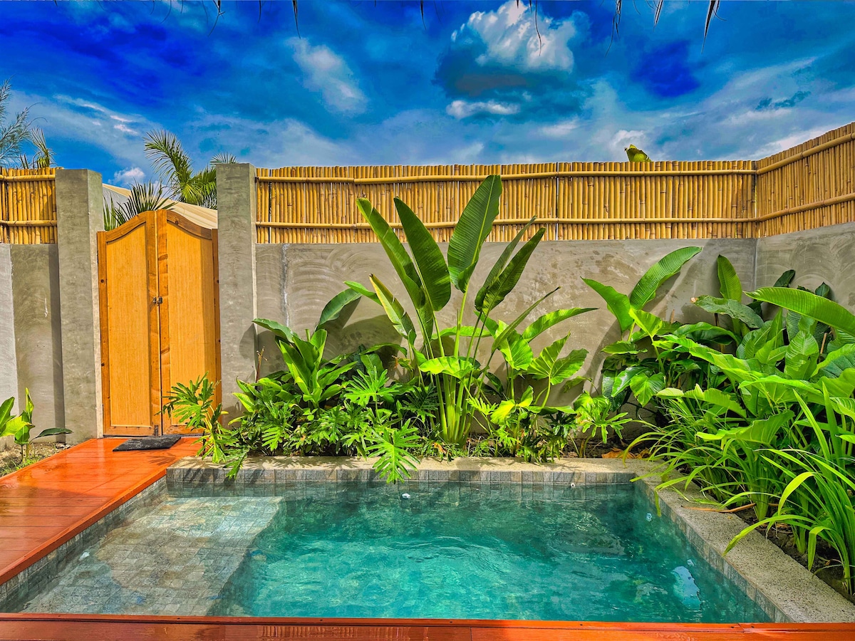 A serene infinity pool is framed by lush tropical plants and a bamboo fence, providing a secluded environment. The water reflects the bright blue sky, with a wooden deck surrounding the pool. Spacious greenery enhances the tranquil setting, inviting relaxation.