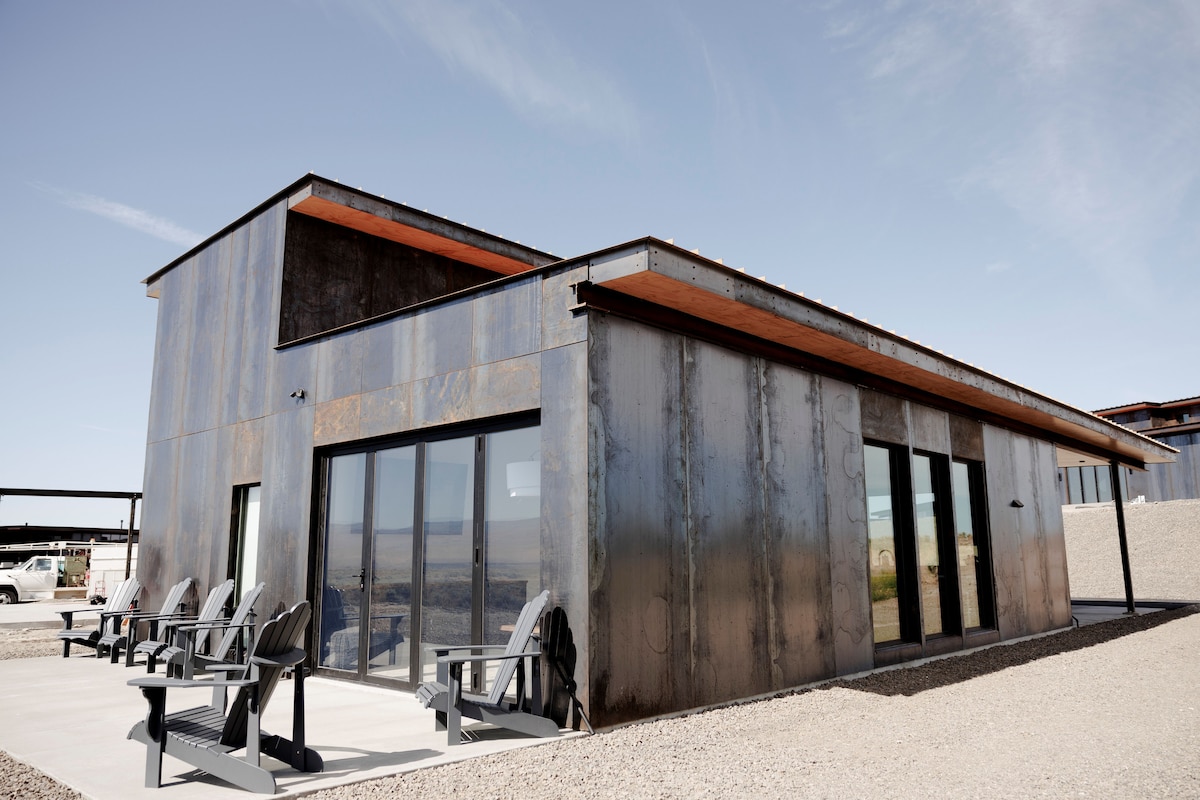 A modern building features a sleek, metallic exterior with large glass doors that provide ample natural light. Outdoor Adirondack chairs are arranged on a concrete patio, offering a comfortable space to enjoy the surrounding landscape.