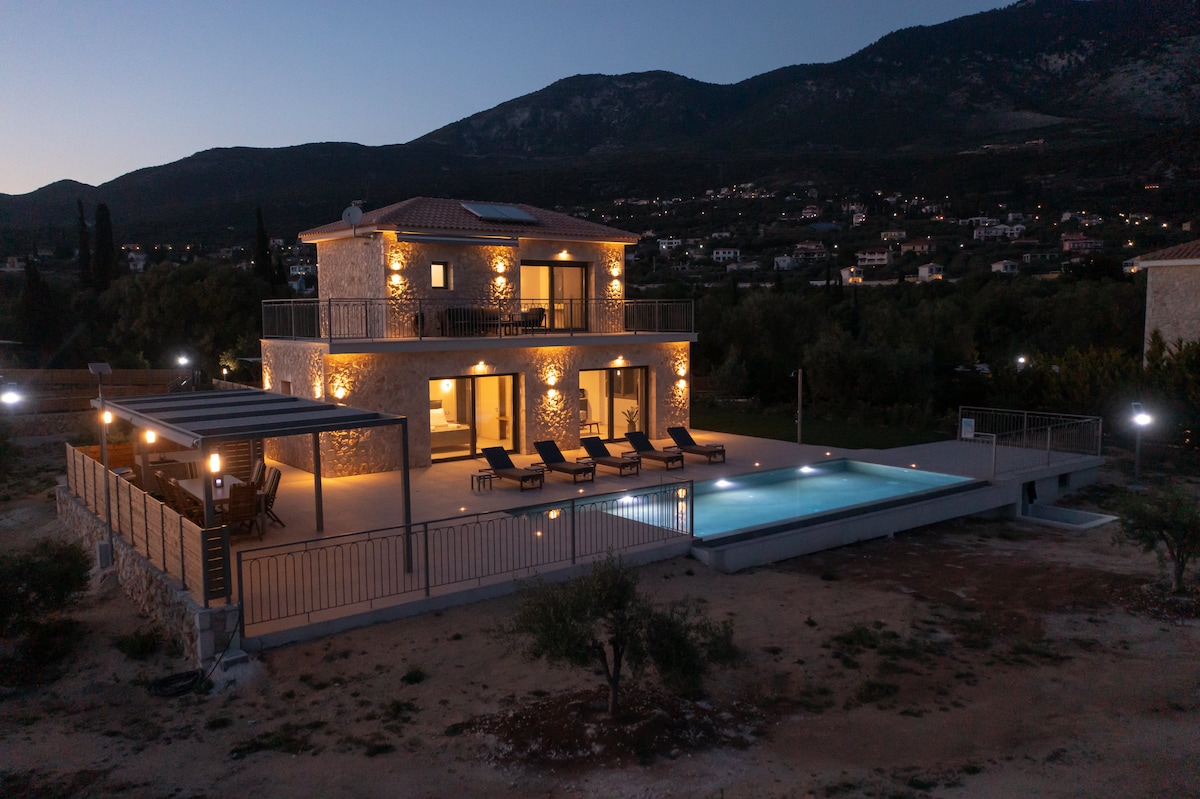 A stone villa is situated in a tranquil outdoor setting, illuminated by soft exterior lighting. The multi-level structure features a spacious terrace and infinity pool, with lounge chairs arranged nearby. Scenic mountains are visible in the background, complementing the serene atmosphere.