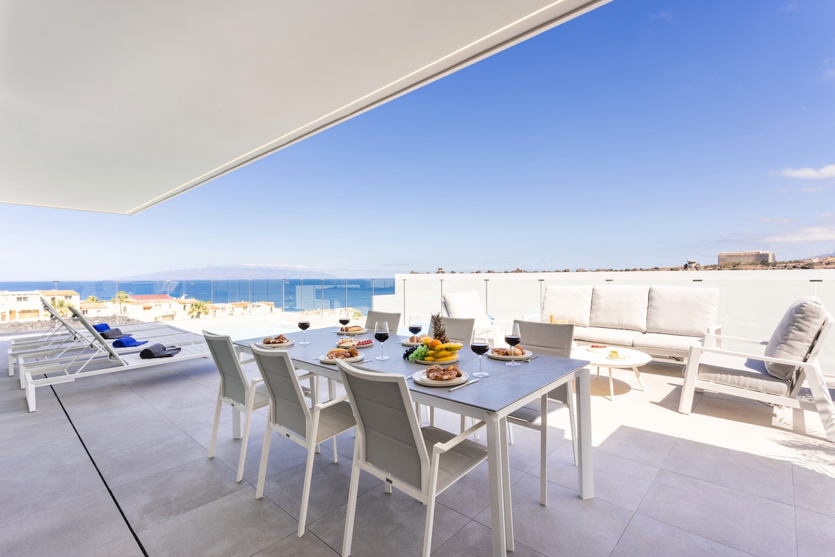 An outdoor dining area is featured on a terrace, showcasing a long table set with plates and glasses, overlooking ocean views. Comfortable seating is arranged nearby, and sun loungers are positioned for relaxation. Clear skies and distant islands enhance the serene atmosphere.