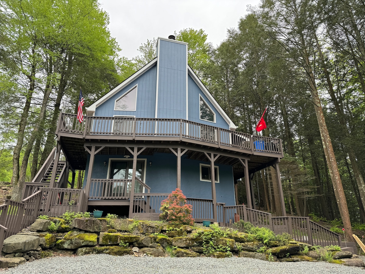A charming blue house is set against a backdrop of tall trees, showcasing a spacious wraparound porch. Flags are displayed prominently by the entrance, and stone landscaping adds a natural element to the property. Steps lead up to the main entrance.