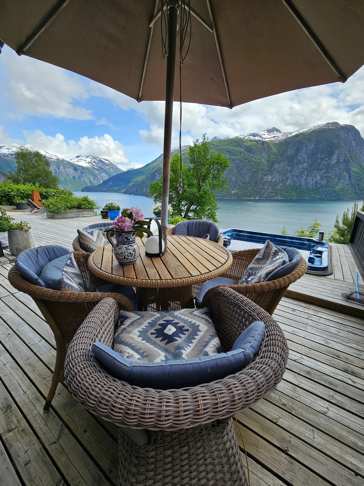 A wooden deck features a round table surrounded by four comfortable wicker chairs, each with soft cushions. An umbrella provides shade, and a scenic view of mountains and water is visible in the background, enhancing the outdoor dining experience.