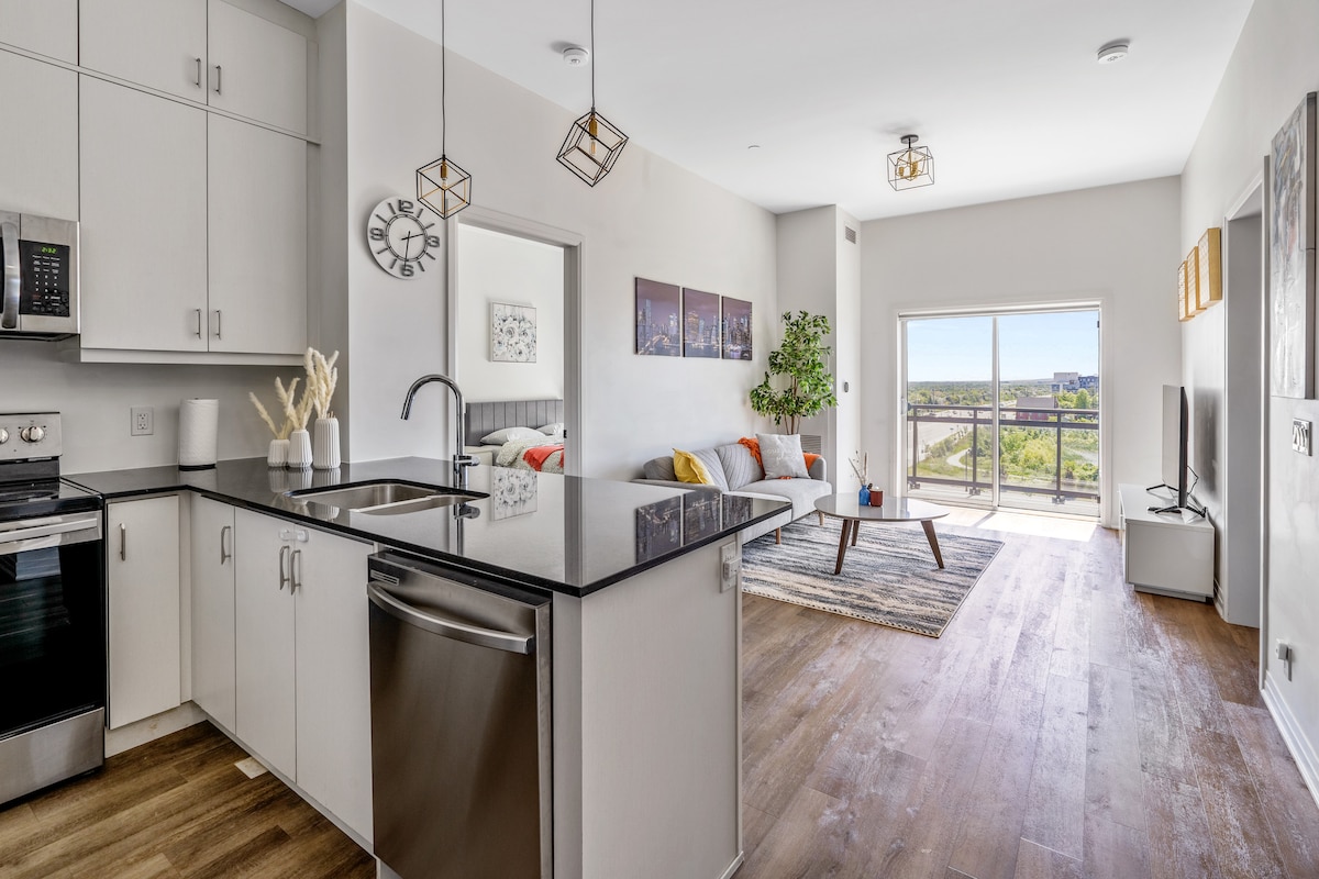 An open living area and kitchen are depicted, featuring modern appliances and a dark countertop. A comfortable seating arrangement with a low table is present, complemented by plants. Natural light flows in through a sliding door, offering a view of the outdoor scenery.