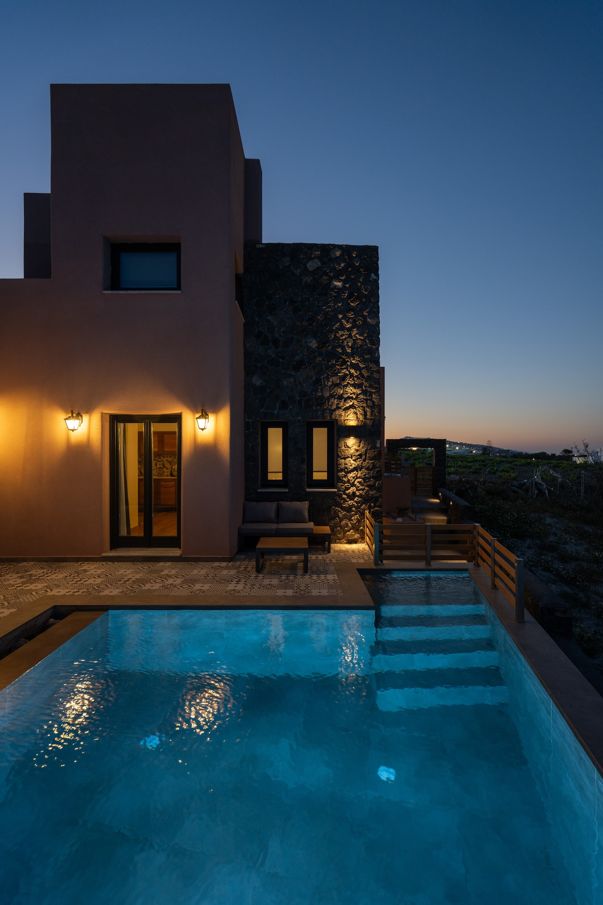 An elegant evening view of the villa is presented, featuring a private swimming pool illuminated softly. The exterior walls, combining smooth and textured surfaces, guide the eye toward the entryway, which is flanked by outdoor seating. A serene sky transitions from twilight to night.