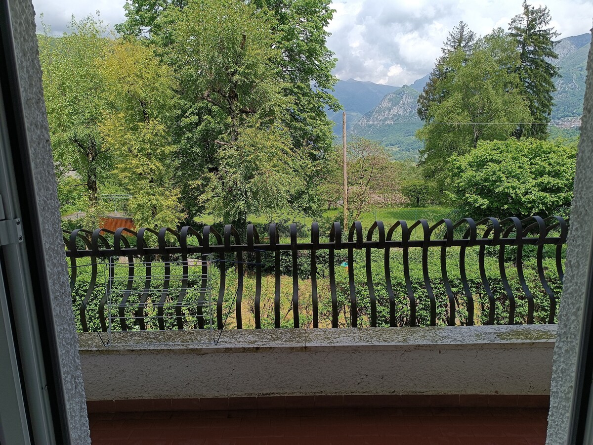 A view from a balcony is presented, showcasing lush greenery and trees against a backdrop of mountains. A decorative black railing frames the scene, offering a sense of openness and connection to the natural surroundings.