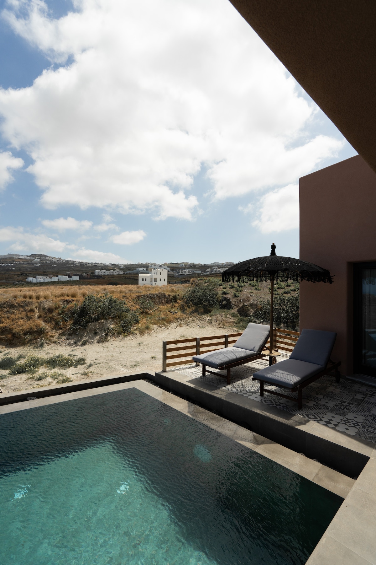 A private swimming pool features a serene blue surface, complemented by two sunbeds with a black umbrella nearby. The surrounding landscape includes gently rolling hills under a cloudy sky, providing a tranquil outdoor setting.