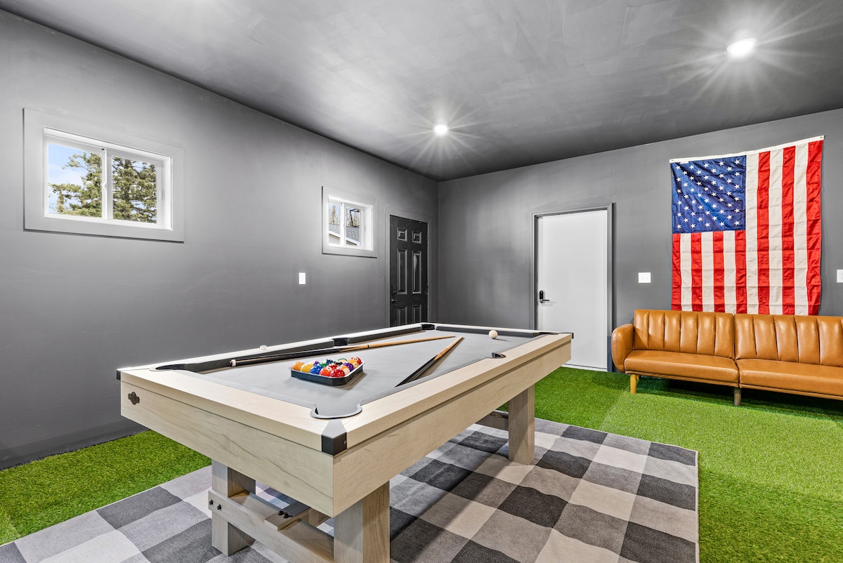 A game room is showcased with a billiards table positioned centrally. A tan couch is placed nearby against a charcoal grey wall, while a large American flag decorates another wall. Two windows provide natural light, and a checkered rug adds texture to the floor.