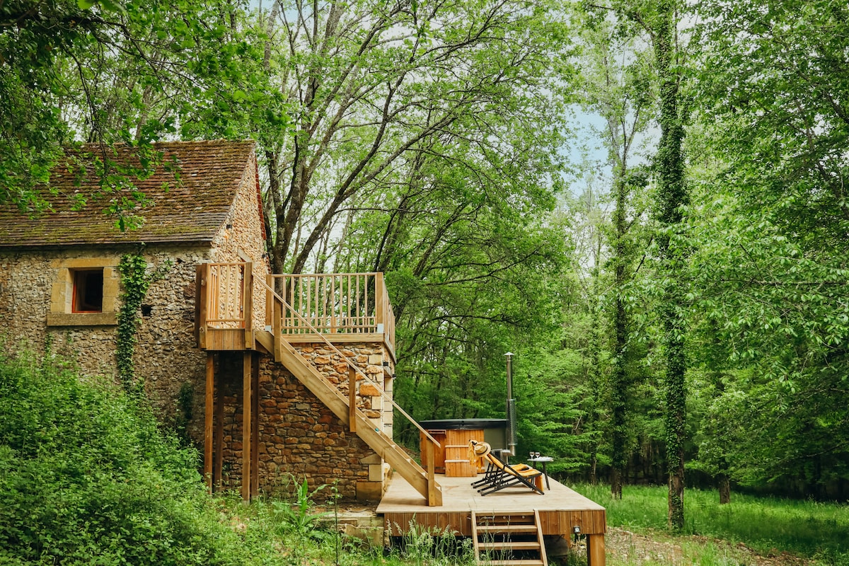 A rustic stone structure is set amidst lush green trees, featuring a wooden deck and stairs leading to an elevated entrance. A hot tub is situated nearby, surrounded by nature, providing a serene outdoor space for relaxation.