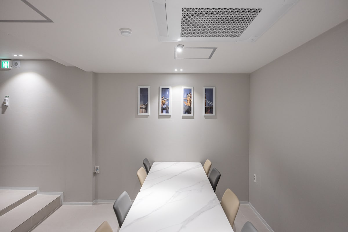 A modern dining area features a long marble table surrounded by six chairs in neutral tones. Four framed photographs are displayed on the light gray wall, adding subtle character to the space. Soft lighting enhances the clean lines of the room.