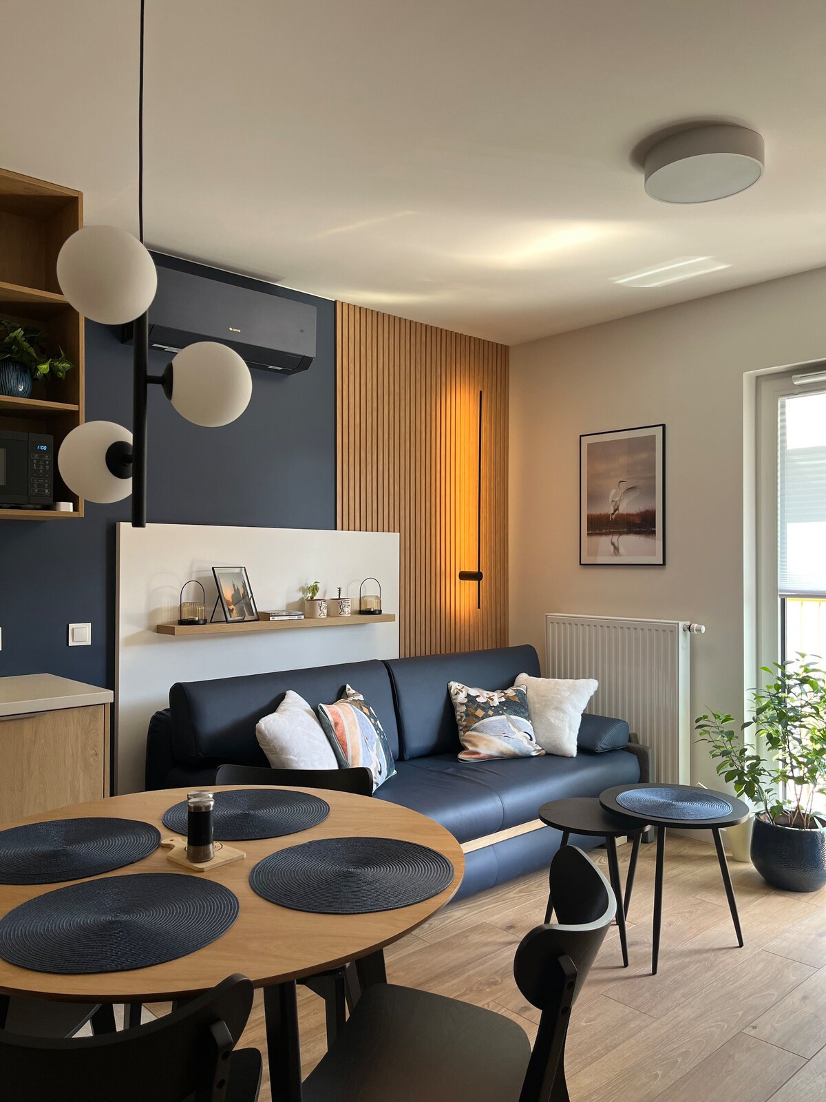 A cozy living space features a navy sofa adorned with decorative pillows and a small round table set with dark placemats. Natural light filters through a nearby window, highlighting wooden accents on the wall and a small green plant in the corner.