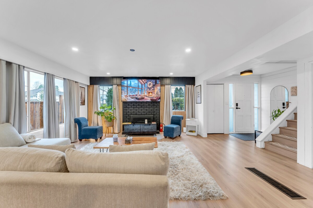 A spacious living area features a cozy arrangement of light-colored couches and accent chairs, complemented by a large, soft area rug. A modern fireplace is centrally positioned, with a flat-screen TV above. Natural light streams in through large windows, enhancing the inviting atmosphere.